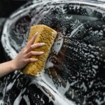 Home A hand scrubs a car window with a sponge, covered in soap and foam, at a car wash.