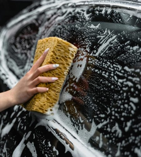 A hand scrubs a car window with a sponge, covered in soap and foam, at a car wash.